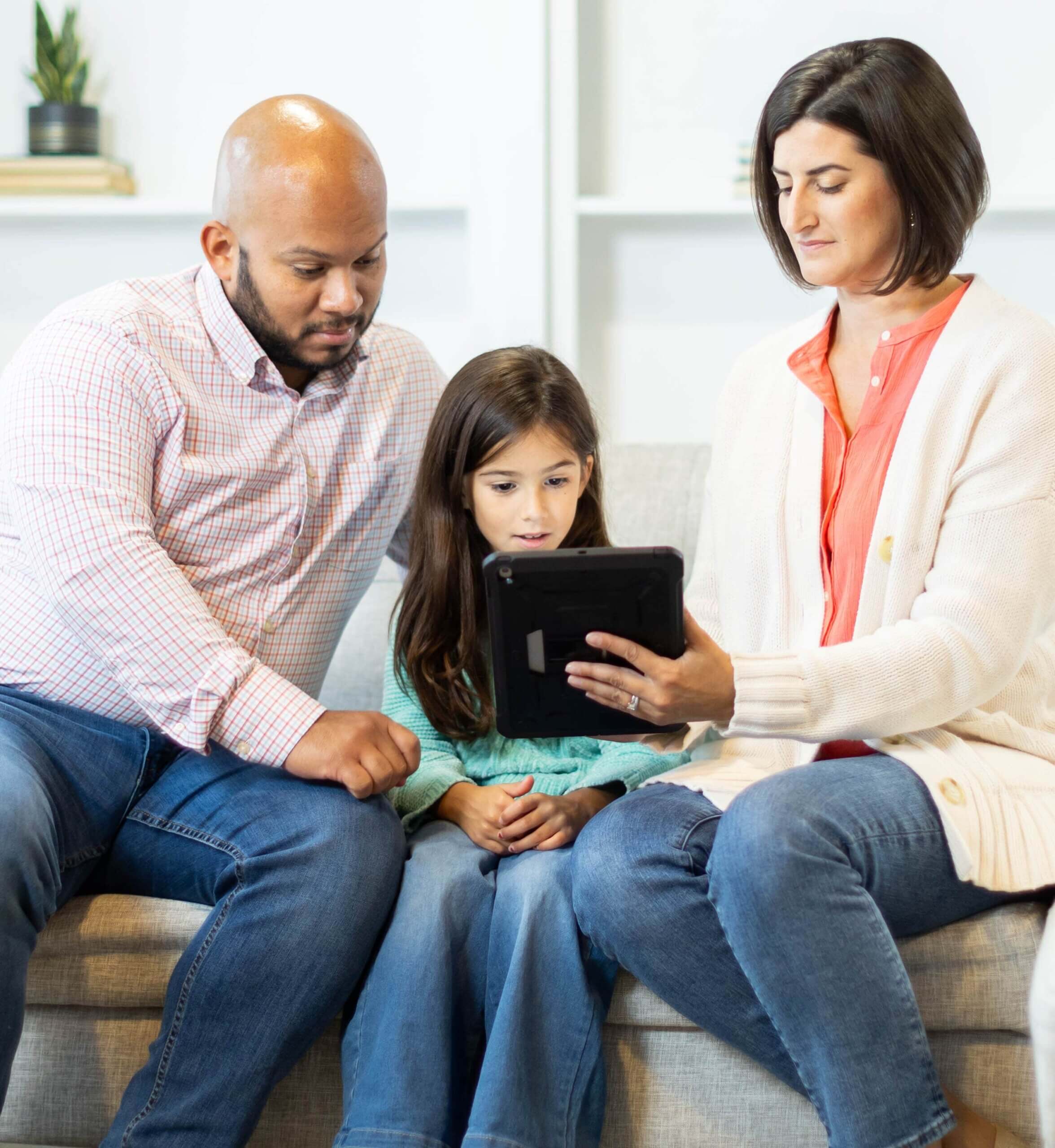 Parents sitting on either side of their child with the mother holding a tablet up to their daughter.