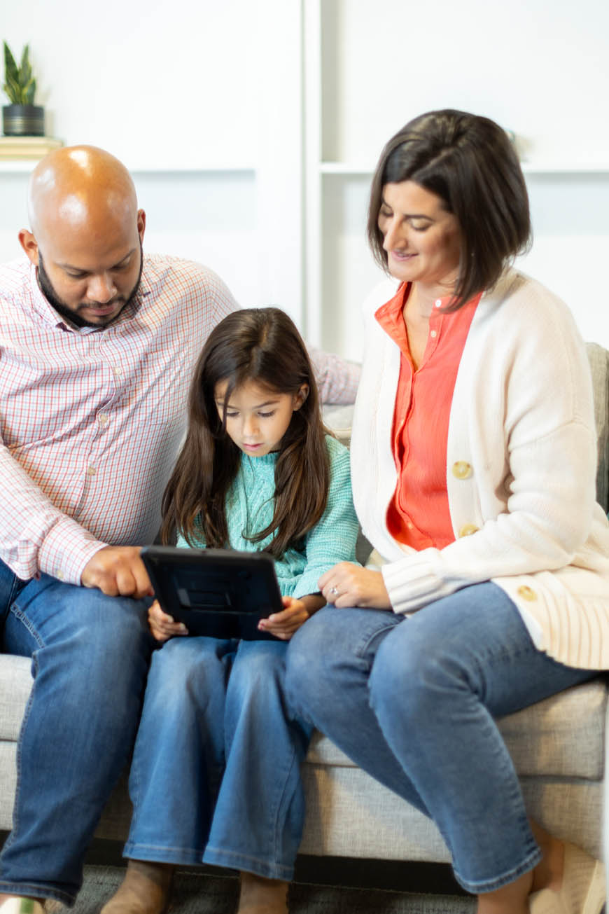 Parents sitting on either side of their child with the mother holding a tablet up to their daughter.